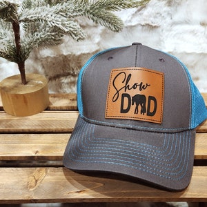 May include: A gray trucker hat with a blue mesh back and brim. The hat features a brown leather patch with the words "Show Dad" and a horse silhouette in black. The hat is sitting on a wooden surface.