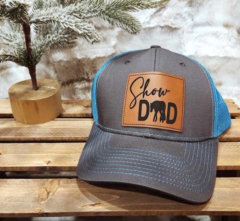May include: A gray trucker hat with a blue mesh back and brim. The hat features a brown leather patch with the words "Show Dad" and a horse silhouette in black. The hat is sitting on a wooden surface.