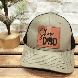 May include: A khaki and black trucker hat with a leather patch. The patch features the words "Show Dad" in a script font and block letters, with a horse silhouette. The hat has a mesh back and a curved brim.