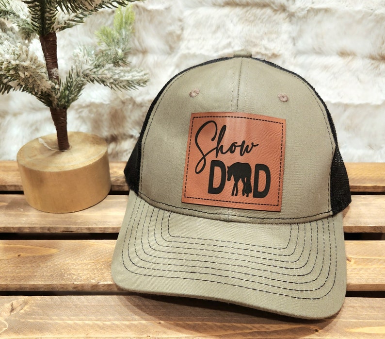 May include: A khaki and black trucker hat with a leather patch. The patch features the words "Show Dad" in a script font and block letters, with a horse silhouette. The hat has a mesh back and a curved brim.