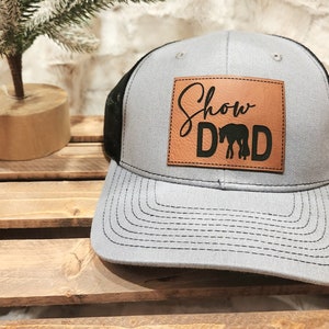 May include: Gray trucker hat with a black mesh back and a leather patch. The patch features the words "Show Dad" above a silhouette of a horse. The hat has a curved brim and a snap closure.