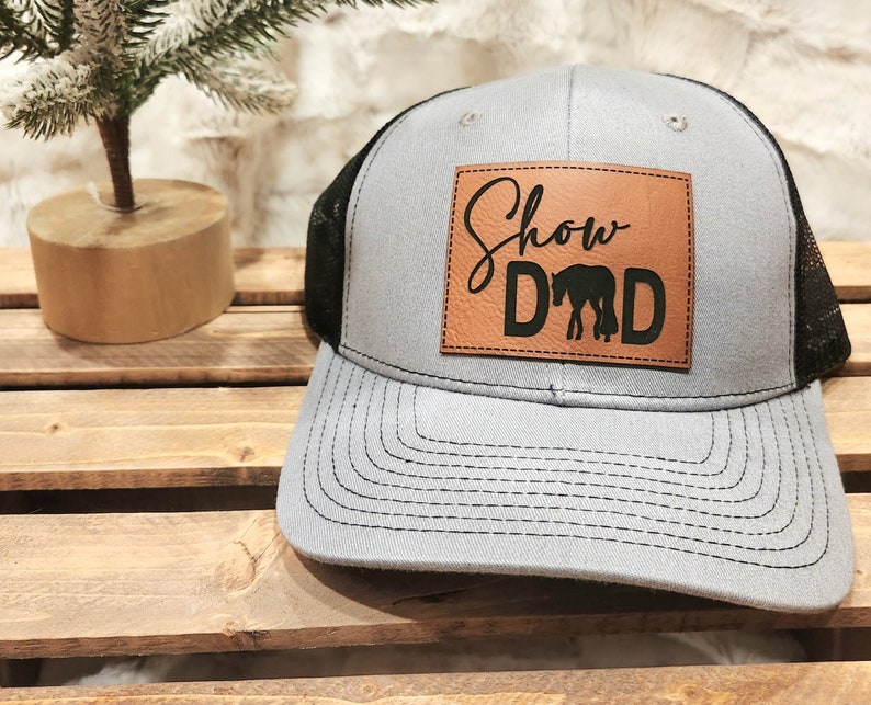 May include: Gray trucker hat with a black mesh back and a leather patch. The patch features the words "Show Dad" above a silhouette of a horse. The hat has a curved brim and a snap closure.