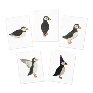 May include: Five white cards each feature a watercolor illustration of a puffin. The puffins are depicted in various poses, with black and white feathers, orange feet, and orange and yellow beaks. One puffin wears a purple party hat.