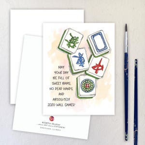 Peut inclure: Carte de v&oelig;ux avec une illustration &agrave; l'aquarelle de tuiles de Mahjong vertes, bleues et rouges. Le texte dit : "Que votre journ&eacute;e soit pleine de doux bams, pas de mains mortes et absolument z&eacute;ro jeu de mur !" Deux pinceaux sont &agrave; droite.