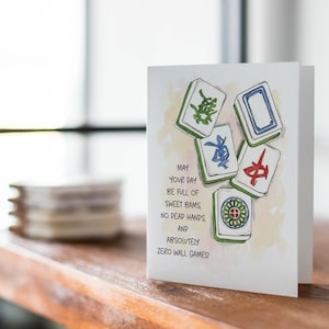 Peut inclure: Une carte de v&oelig;ux blanche avec des illustrations &agrave; l'aquarelle de tuiles de Mahjong. La carte contient le texte : "MAY YOUR DAY BE FULL OF SWEET BAMS, NO DEAD HANDS, AND ABSOLUTELY ZERO WALL GAMES !" La carte est sur une surface en bois.