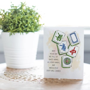 Peut inclure: Carte de v&oelig;ux avec des illustrations &agrave; l'aquarelle de tuiles de Mahjong. La carte indique : "Que votre journ&eacute;e soit pleine de bams doux, pas de mains mortes et absolument aucun jeu de mur !" Un pot de plante blanche est en arri&egrave;re-plan.