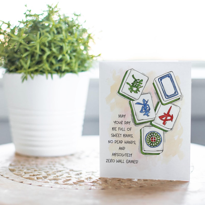 Peut inclure: Carte de v&oelig;ux avec des illustrations &agrave; l'aquarelle de tuiles de Mahjong. La carte indique : "Que votre journ&eacute;e soit pleine de bams doux, pas de mains mortes et absolument aucun jeu de mur !" Un pot de plante blanche est en arri&egrave;re-plan.