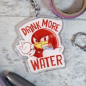 May include: A red and white keychain featuring a cartoon character holding a glass of water. The text "Drink More Water" is written in red letters.