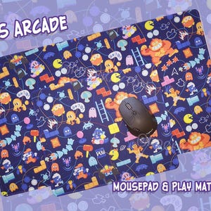 May include: A navy blue mousepad and play mat with a colorful 80s arcade game design. The mat features various game characters and elements, with the text "80S ARCADE" and "MOUSEPAD & PLAY MAT". A black computer mouse is on the mat.