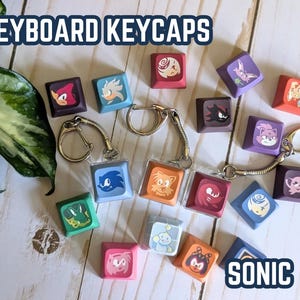 May include: A collection of square keyboard keycaps featuring various colorful character illustrations from the Sonic franchise. Three of the keycaps are attached to metal keychains. The text KEYBOARD KEYCAPS and SONIC is visible.