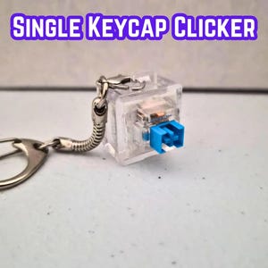 May include: A clear plastic mechanical keyboard switch keychain with a blue stem, attached to a silver metal split ring by a flexible coiled spring connector. The text SINGLE KEYCAP CLICKER is displayed in purple letters above the object.