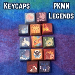 May include: A collection of square keycaps featuring various colorful Pokémon designs. The keycaps are arranged on a blue and purple background, with the words "KEYCAPS" and "PKMN LEGENDS" in white text.