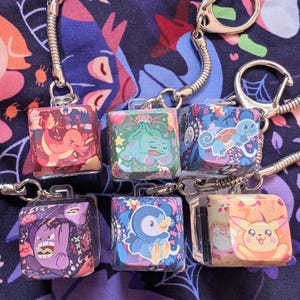 May include: A collection of six cube-shaped keychains featuring colorful cartoon character designs. Each keychain has a silver-tone metal clasp and a small handle. The characters are set against a floral and geometric patterned background.