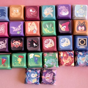 May include: A collection of colorful square keycaps featuring various cartoon character designs. The keycaps are arranged in rows and columns against a pink background. Each keycap displays a different character in vibrant colors, creating a visually appealing and playful aesthetic.