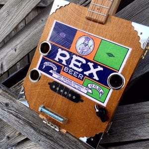 May include: A handmade cigar box guitar with a wooden body and a Rex Beer label. The guitar features a bolt bridge, metal corner protectors, and a black pickup. The label includes the text "REX BEER" and "12 FL.OZ."
