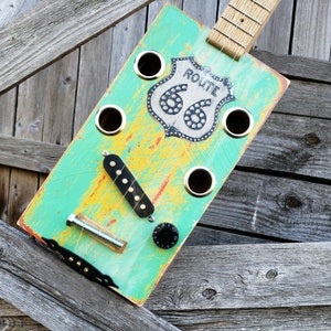 May include: A teal and yellow painted cigar box guitar with a Route 66 design and a black pickup. The guitar has a silver tuning knob and a silver bolt.