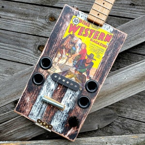 May include: A cigar box guitar with a black and white distressed finish. The guitar has a single pickup and a bridge with a metal bar. The guitar is made from a repurposed cigar box with a vintage Western magazine cover featuring cowboys and horses. The magazine cover reads "10 Dime Western Magazine".