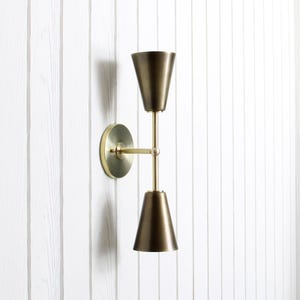 May include: A brass wall sconce with a conical shade at the top and bottom, connected by a straight rod. The sconce is mounted on a circular base and is against a white, vertical-slat wall.