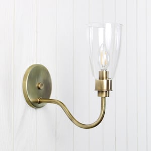 May include: A wall-mounted sconce with a clear glass shade and a brass finish. The sconce features a curved arm and a round backplate. The light bulb is visible inside the glass shade. The fixture is designed for indoor use.
