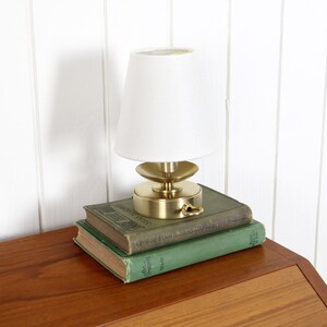 May include: A small table lamp with a brushed brass base and a white conical shade, resting on top of two stacked green hardcover books. The lamp features a circular brass platform, a central stem, and a small ring pull on the side of the base.