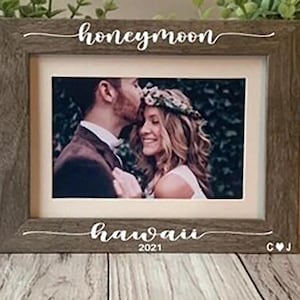May include: A brown wooden picture frame with a white inscription that reads "honeymoon hawaii 2021" and a photo of a couple kissing. The frame also has the initials "C & J" on the bottom right corner.