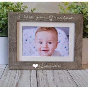 May include: A framed photograph with a smiling baby. The frame is a weathered gray color with the words "I love you Grandma" at the top and the name "James" at the bottom, with a heart symbol. The photo is surrounded by a white mat.