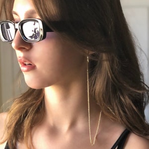Peut inclure: Une femme portant des lunettes de soleil à monture noire avec une chaîne dorée attachée aux lunettes et qui pend autour de son cou.
