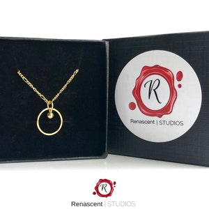 May include: A gold necklace with a small circle pendant and a single bead. The necklace is in a black velvet box with a white label that says "Renascent | STUDIOS".