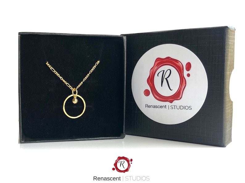 May include: A gold necklace with a small circle pendant and a single bead. The necklace is in a black velvet box with a white label that says "Renascent | STUDIOS".