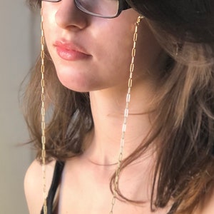 May include: A gold chain with small, rectangular links hangs from the ears of a person wearing glasses. The chain is a glasses chain, designed to keep glasses from falling off.
