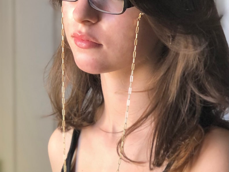 May include: A gold chain with small, rectangular links hangs from the ears of a person wearing glasses. The chain is a glasses chain, designed to keep glasses from falling off.