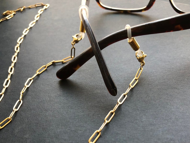 May include: A gold chain with oval links connects to two small gold clasps. The clasps are attached to the arms of a pair of tortoiseshell eyeglasses.