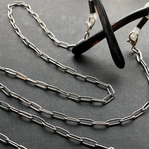 May include: A silver chain with two spring-loaded clips for attaching to glasses. The chain is made of linked oval shapes.