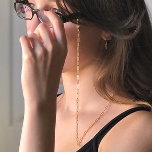 May include: A gold chain with small, rectangular links hangs from the ears of a person wearing glasses. The chain is attached to the glasses with small clips.