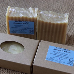 May include: Two bars of handmade Patchouli Delight Shampoo & Body Bar soap, each wrapped in clear plastic and sitting on a brown burlap surface. The soap is a light brown color and has a textured surface. The soap is packaged in a brown cardboard box with a window to show the soap inside. The box has a blue label with the text "Patchouli Delight Shampoo & Body Bar" and a list of ingredients.