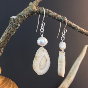 Puede incluir: Un par de pendientes con una perla blanca y una rebanada de asta de ciervo. La asta de ciervo es de color marrón claro y tiene una textura natural y orgánica. Los pendientes cuelgan de ganchos de plata.