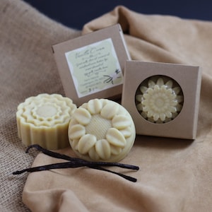May include: Three handmade soap bars in the shape of flowers, with a vanilla cream scent. The soaps are packaged in brown cardboard boxes with a white label that reads "Vanilla Cream with Almond Oil &  Gentleman's Blend by  Soap  &  Candle  Co."