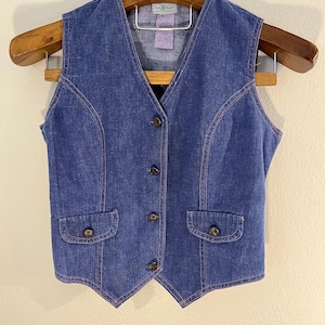 Könnte beinhalten: Eine blaue Jeansweste mit V-Ausschnitt, zwei Vordertaschen und braunen Knöpfen. Die Weste hängt an einem Holzkleiderbügel. Die Nähte sind in einer kontrastierenden beigen Farbe.