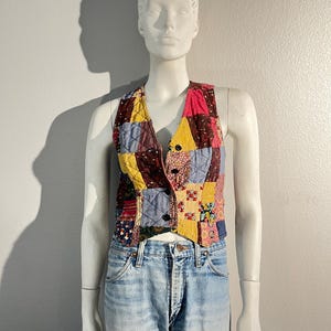 May include: A colourful patchwork vest with a V-neck. The vest is made from various fabric squares in blue, yellow, red, and brown, sewn together in a quilted pattern. It has a button-up front and is displayed on a mannequin.