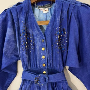 May include: A blue denim dress featuring a V-neckline, gold-tone buttons, and puffed sleeves. The bodice is embellished with clusters of dark blue circular studs. A matching fabric belt with a circular buckle featuring blue studs is included.