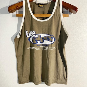Może przedstawiać: Oliwkowy podkoszulek z białym wykończeniem i napisem "Lee Pipes" w niebiesko-białym wzorze. Podkoszulek wisi na drewnianym wieszaku. Dodatkowy tekst to "MADE ORIGINAL" i "LOGONSION IS A REGISTERED TRADEMARK OF STYLING MR. LEE JEANS AND CASUAL WEAR ORIGINAL FITTING."