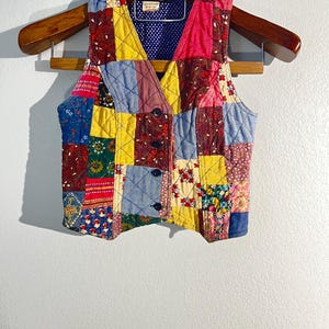 May include: A patchwork vest with a V-neck design, featuring a variety of colorful fabric squares. The vest is quilted and has a blue lining. It is hanging on a wooden hanger.