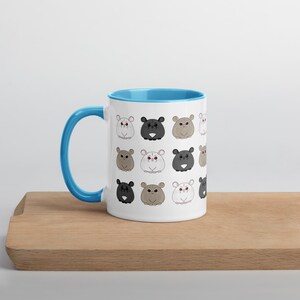 May include: White ceramic mug with a light blue interior and handle. The mug is decorated with a repeating pattern of cartoon bear faces in various colors, including black, gray, and white. The mug sits on a wooden surface.