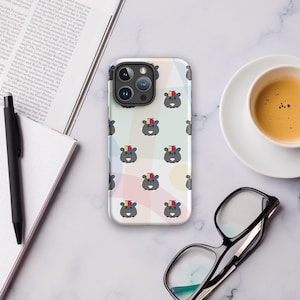 May include: A smartphone case featuring a repeating pattern of cartoon hippos wearing rainbow-striped hats. The case has a white background with pastel color blocks. A cup of coffee, glasses, and a notebook are also visible.