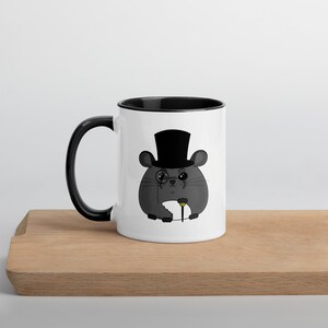 May include: A white ceramic mug with a black interior and handle. The mug features a cartoon illustration of a gray mouse wearing a black top hat, monocle, and holding a cane. The mug is sitting on a wooden surface.