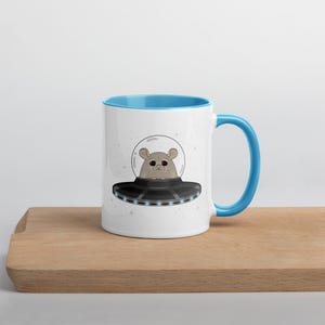 May include: White ceramic mug with a blue interior and handle. The mug features a cartoon illustration of a hamster in a spaceship. The spaceship is black with a gray rim and a clear bubble. The background is white with small star illustrations.