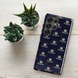 May include: A dark blue phone case featuring a repeating pattern of cartoon bears in UFOs against a starry background. The case has a glossy finish and a camera module with multiple lenses. Two small potted succulents are in the background.