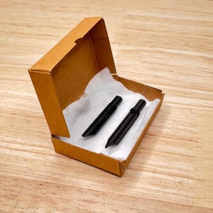 May include: Two black calligraphy pen nibs are presented in an open, small cardboard box, lined with white padding. The box is light brown and rests on a wooden surface. The nibs are angled and appear unused.