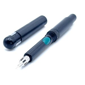 May include: A black fountain pen with a silver nib and a detached cap. The pen has a teal-colored button and a textured grip. The pen is lying on a white surface.