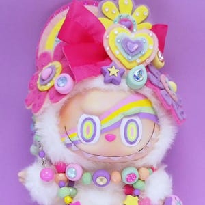 May include: A whimsical plush doll with a white furry body and a smiling face. It is adorned with a rainbow-striped forehead, a pink bow, and a colorful beaded necklace. The doll is decorated with hearts, stars, and flowers, set against a purple background.